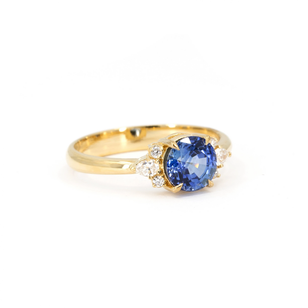 Yellow gold engagement ring featuring a round blue sapphire center stone with pear and round diamond side accents. Designed by Bena Jewelry, handcrafted in Montreal.