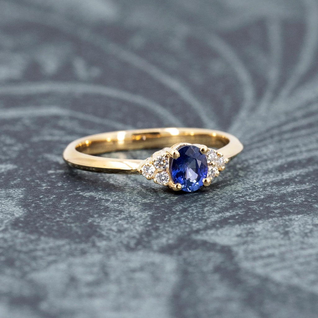 A classic yellow gold engagement ring handmade in Montreal by independent local jewelry brand Ruby Mardi is seen photographed on a romantic dark green background. This wedding ring shows a central oval blue royal sapphire and symmetrical natural diamond accents.