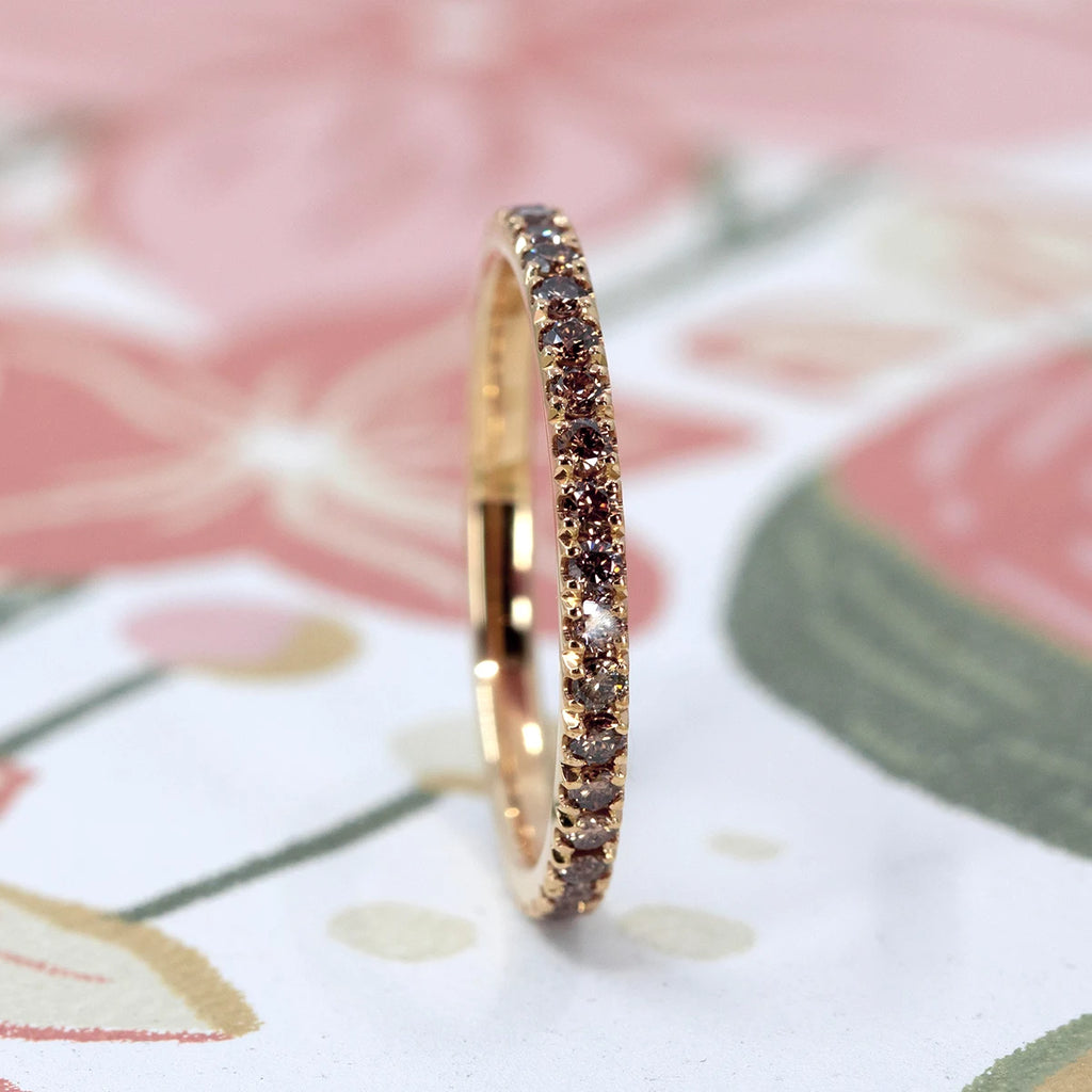 Brown diamond eternity band in yellow gold photographed upright on a floral background. Fine pavé setting with warm-toned diamonds creating a delicate, continuous line of light.