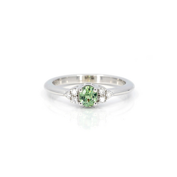 A classic white gold engagement ring is photographed on a white background. The ring features 6 round brilliant diamonds as accents on the sides of a central oval green gemstone (a demantoid garnet). This bridal ring was handmade in Montreal.