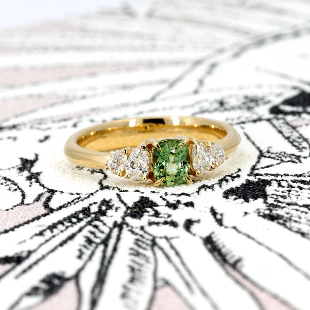 A handcrafted gold engagement ring by Ruby Mardi, featuring a cushion-cut green sapphire at the center, flanked by pear-shaped diamonds. Made in Montreal with ethical materials, this elegant yet modern design is displayed on an artistic black and white background, enhancing the brilliance of the gemstones and the warmth of the gold setting.







