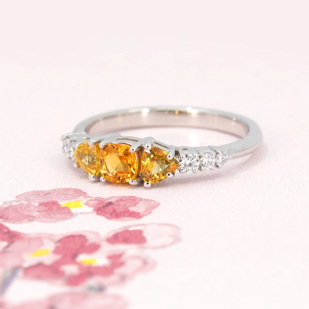 A colorful classic piece of jewelry featuring 3 orange sapphires and 5 extra brilliant white diamonds is photographed on a watercolor image of flowers. This ring was designed by Ruby Mardi, an independent jewelry store in Montreal, Canada.