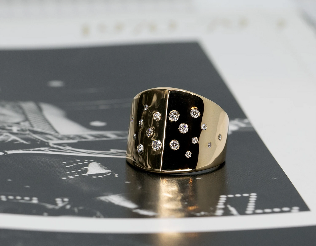 Wide yellow gold statement ring with an asymmetric polished surface, featuring scattered round white diamonds in a modern, edgy design. Created by Bena Jewelry, Montreal, Canada.