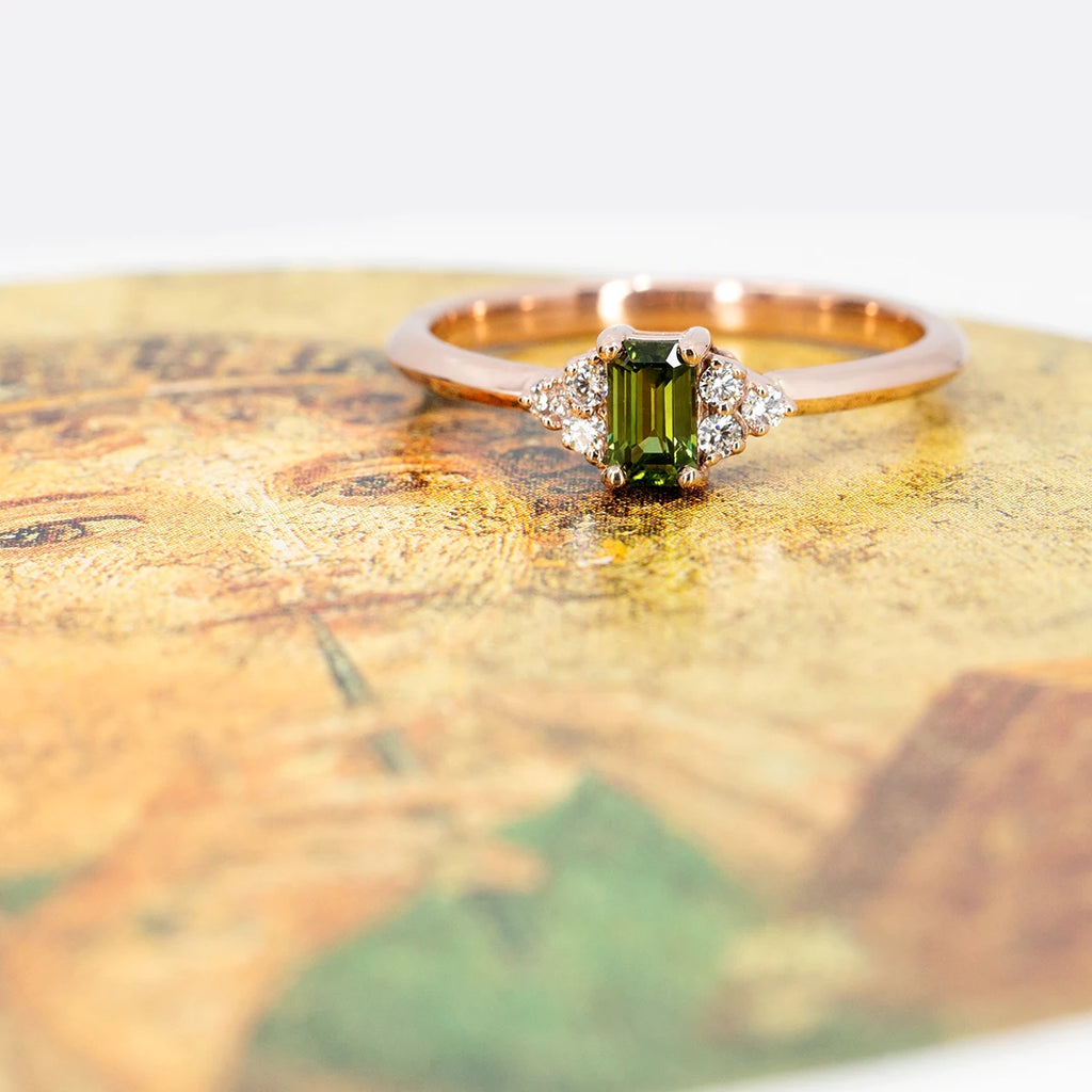 Alternative engagement ring on the dainty side is photographed on an old painting showing a flute player. The ring is in rose gold and features a rectangular green sapphire, as well as three diamonds on each side. Custom made jewel. Available at Ruby Mardi in Montreal's Little Italy.