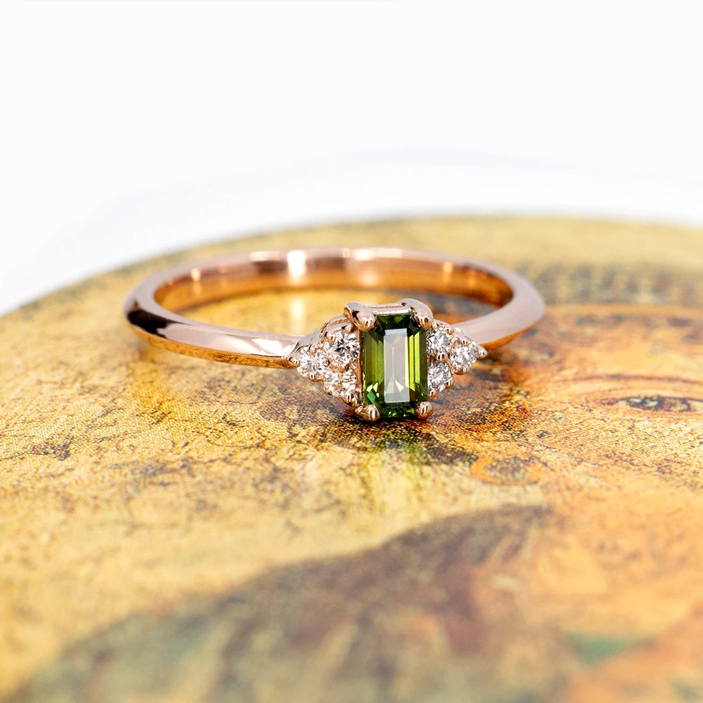 A rose gold ring with a central rectangular green emerald and round brilliant diamond accents is photographed on a renaissance painting. This fine jewelry piece was handcrafted in Montreal by local independent brand Ruby Mardi.