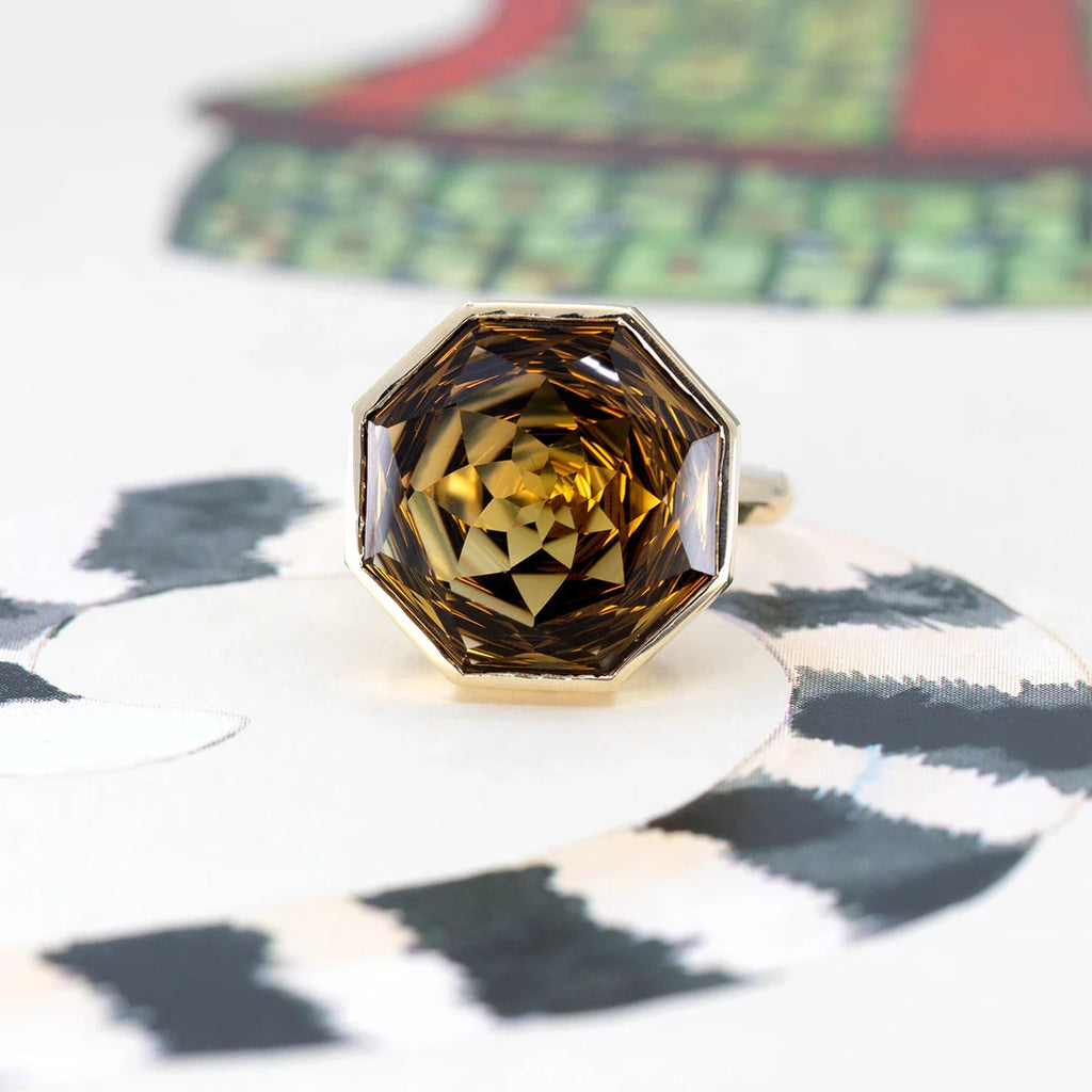 A front view of a large, funky yellow gold ring with lots of style is photographed against a colorful abstract background. The ring features a huge dark natural citrine in a bezel setting. Available at the independent Canadian jewelry store Ruby Mardi.