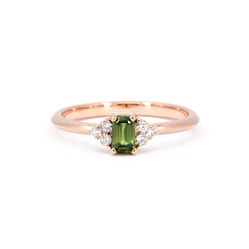 Dainty rose gold engagement ring with a central emerald shape green sapphire and six lab grown diamond accents on each side. The ring was handmade in Montreal and is seen photographed on a white background.