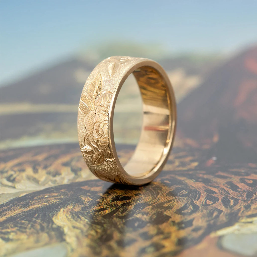 7 mm hand-engraved floral gold band by Rebel & Rogue, featuring detailed peony carvings with a textured stippled background. Solid yellow gold wedding ring photographed on an illustrated surface at Ruby Mardi, Montreal jewelry store.