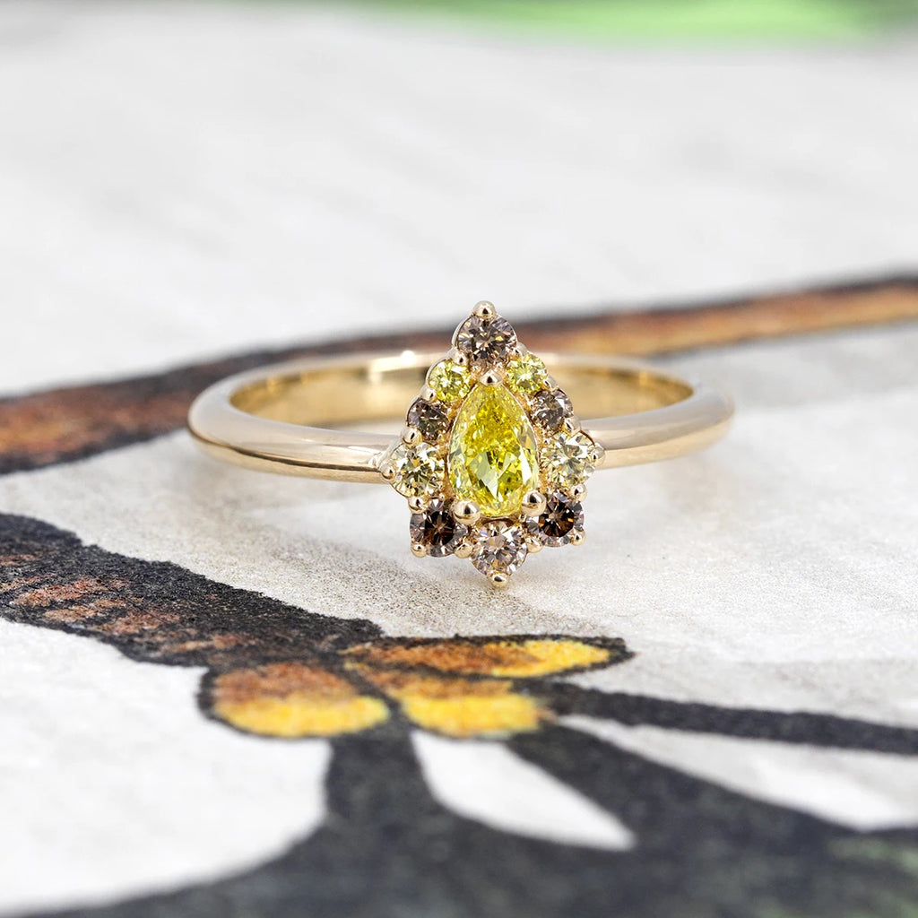 Yellow diamond halo ring with pear-shaped center stone, set in yellow gold with brown and yellow diamonds. Photographed on illustrated background. Ruby Mardi, Montreal.