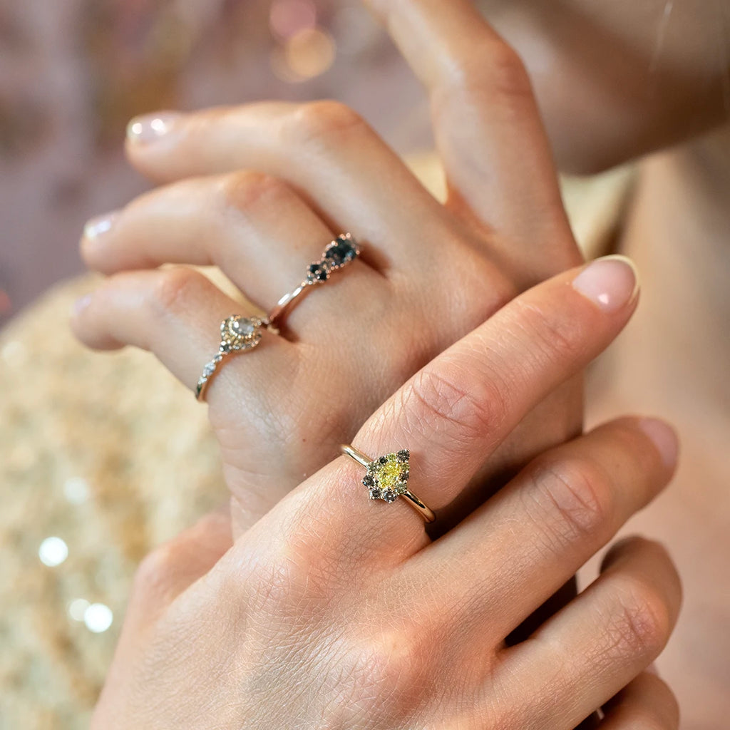 Close-up of a woman’s hands wearing three rings: a yellow gold engagement ring with a pear-shaped yellow diamond halo, a white gold ring with dark and white diamond clusters, and a white gold ring with a round white diamond in a vintage-inspired setting. Designer jewelry by Ruby Mardi, Montreal, Canada.
