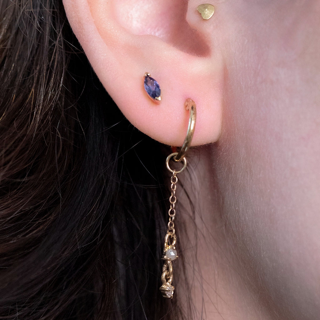 Close-up of a person wearing a natural marquise-cut iolite stud earring in 10k yellow gold, shown on the earlobe next to a gold hoop, for Ruby Mardi in Montreal.