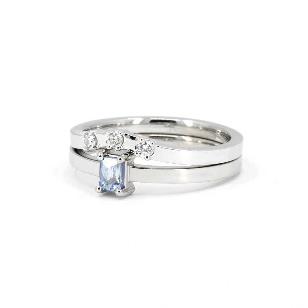 White gold ring set featuring a light blue sapphire baguette solitaire ring paired with a diamond crown band. The solitaire ring showcases a sleek horizontal baguette cut, while the crown band adds brilliance with three round diamonds in a curved design. A modern and refined bridal set crafted by Ruby Mardi in Montreal.