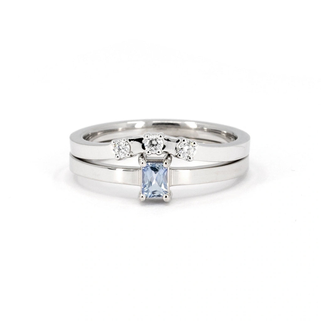 Elegant white gold ring set featuring a light blue sapphire baguette solitaire ring paired with a matching diamond crown band. The solitaire ring showcases a horizontally set baguette-cut sapphire with a soft, icy blue hue, while the crown band adds brilliance with three round diamonds in a curved design. A modern and sophisticated combination crafted by Ruby Mardi in Montreal. Perfect as a bridal set or for stacking.