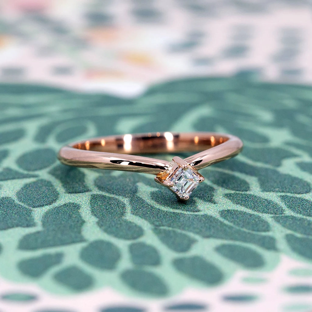 A delicate 14k rose gold ring featuring an Asscher cut diamond set at an angle. The minimalist band has a refined, modern design, photographed against a textured green background with abstract organic patterns. Designed and crafted by Ruby Mardi in Montreal.
