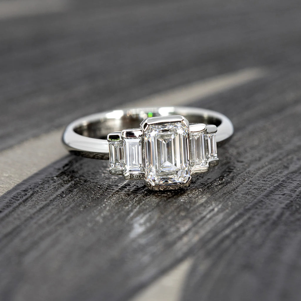 Close-up of a white gold diamond ring with a central emerald-cut diamond and straight baguette diamonds on each side, shown on a textured black surface. This Art Deco-inspired engagement ring is handcrafted in Montreal by Ruby Mardi and features bold symmetry with clean modern lines.