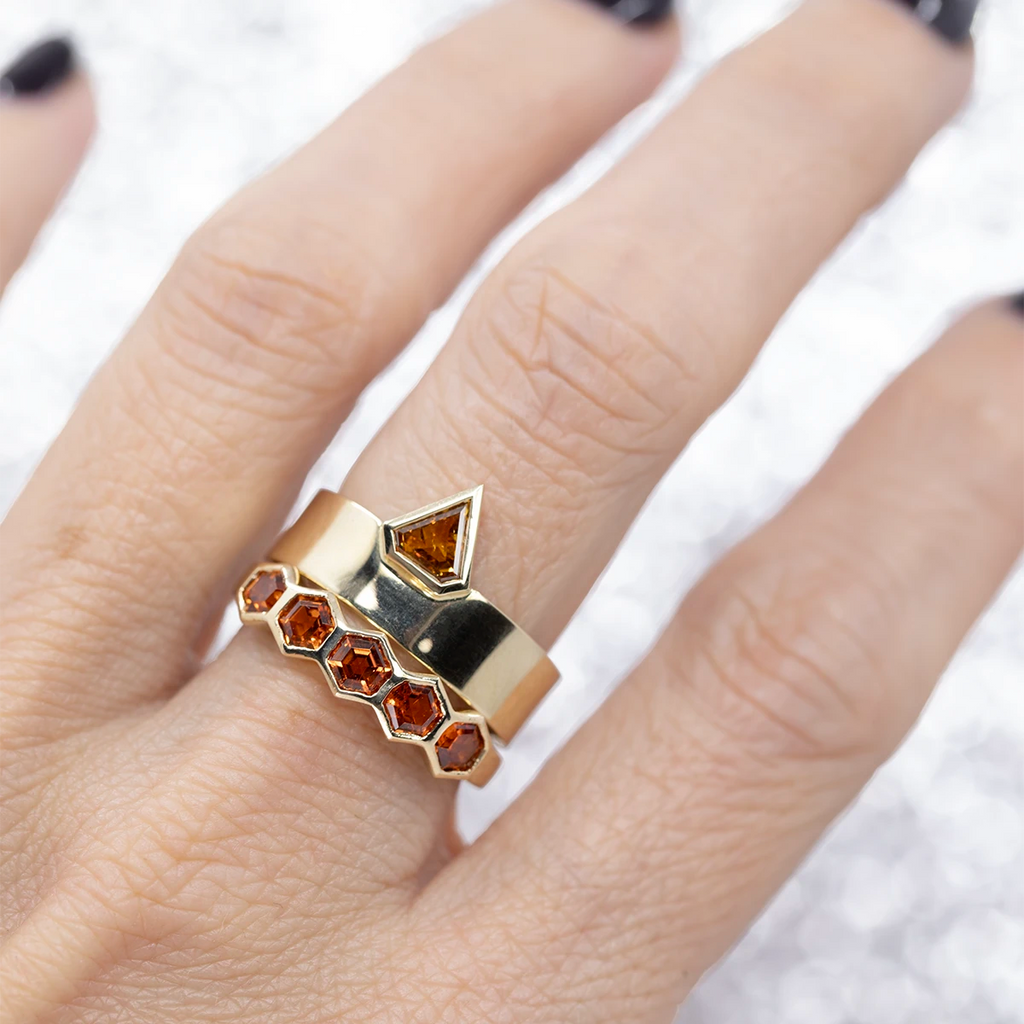 Hand photographed close-up, showing two yellow gold rings with orange gemstones worn on the middle finger. The top ring is a wide gold band topped by a triangular orange diamond, and the bottom ring features several hexagonal-cut orange garnets. Two fine jewels by independent designers available at Ruby Mardi, Montreal, Canada.