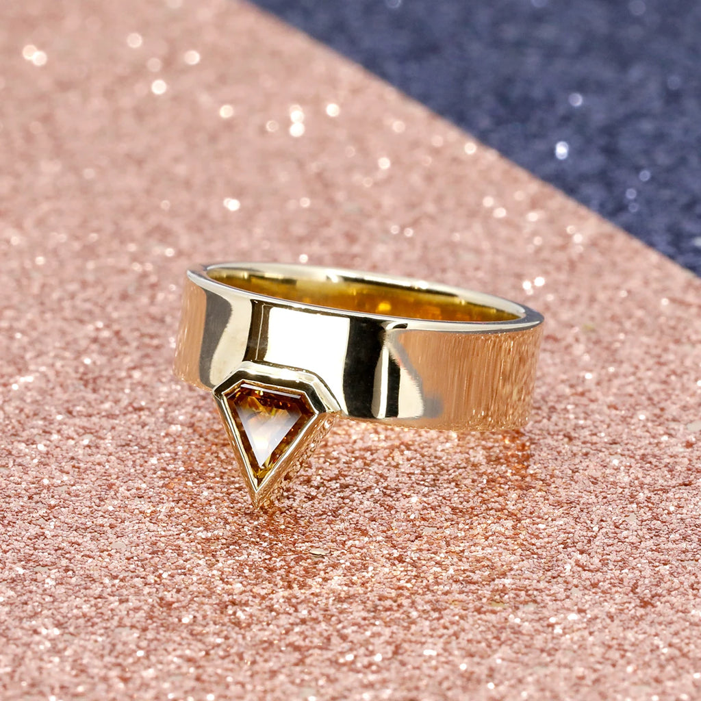 Thick gold band with a pantagonal orange diamond in bezel set is seen photographed on pink glitters. This statement gender neutral ring was designed by independent brand Bena Jewelry and is available at the best jewelry store in Montreal, Ruby Mardi.