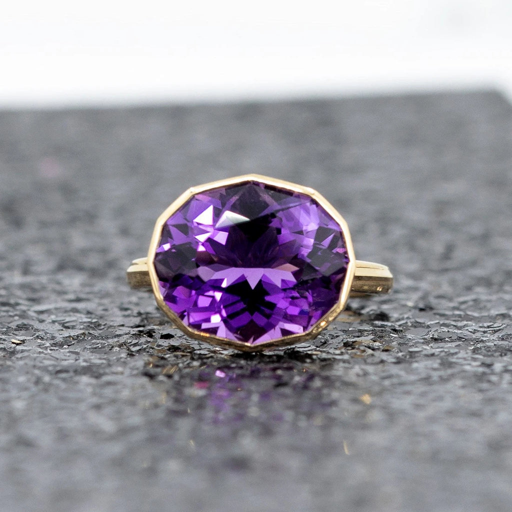 Front view of a big oval genuine amethyst of profound purple color and pink hues bezel set in 14k yellow gold, seen photographed on a black sequins background. This is a creation from Canadian jewelry designer Bena Jewelry.
