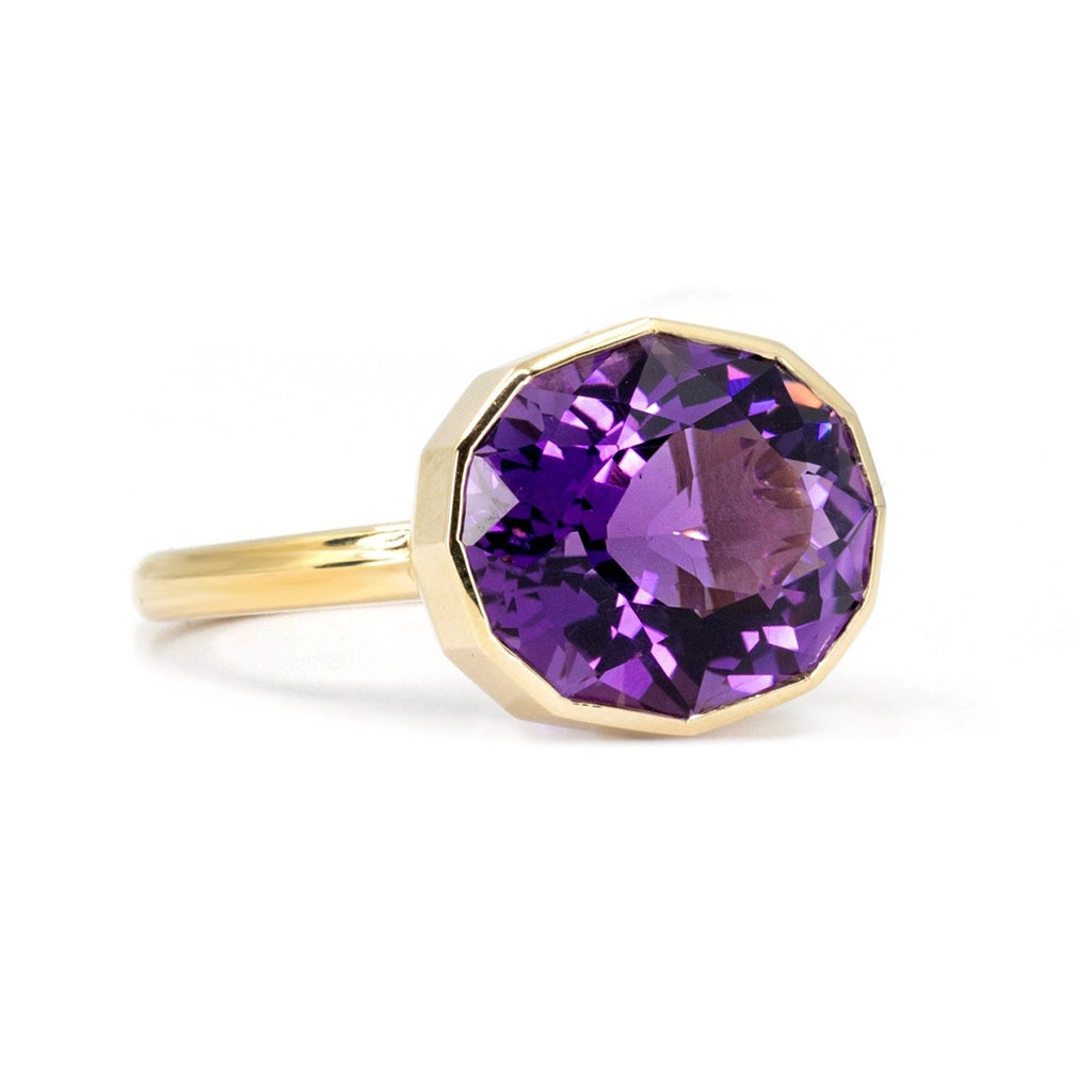 Side view of a big oval shape natural amethyst of profound purple color and pink hues bezel set in 14k yellow gold, seen photographed on a white background. This is a creation from Canadian jewelry designer Bena Jewelry.