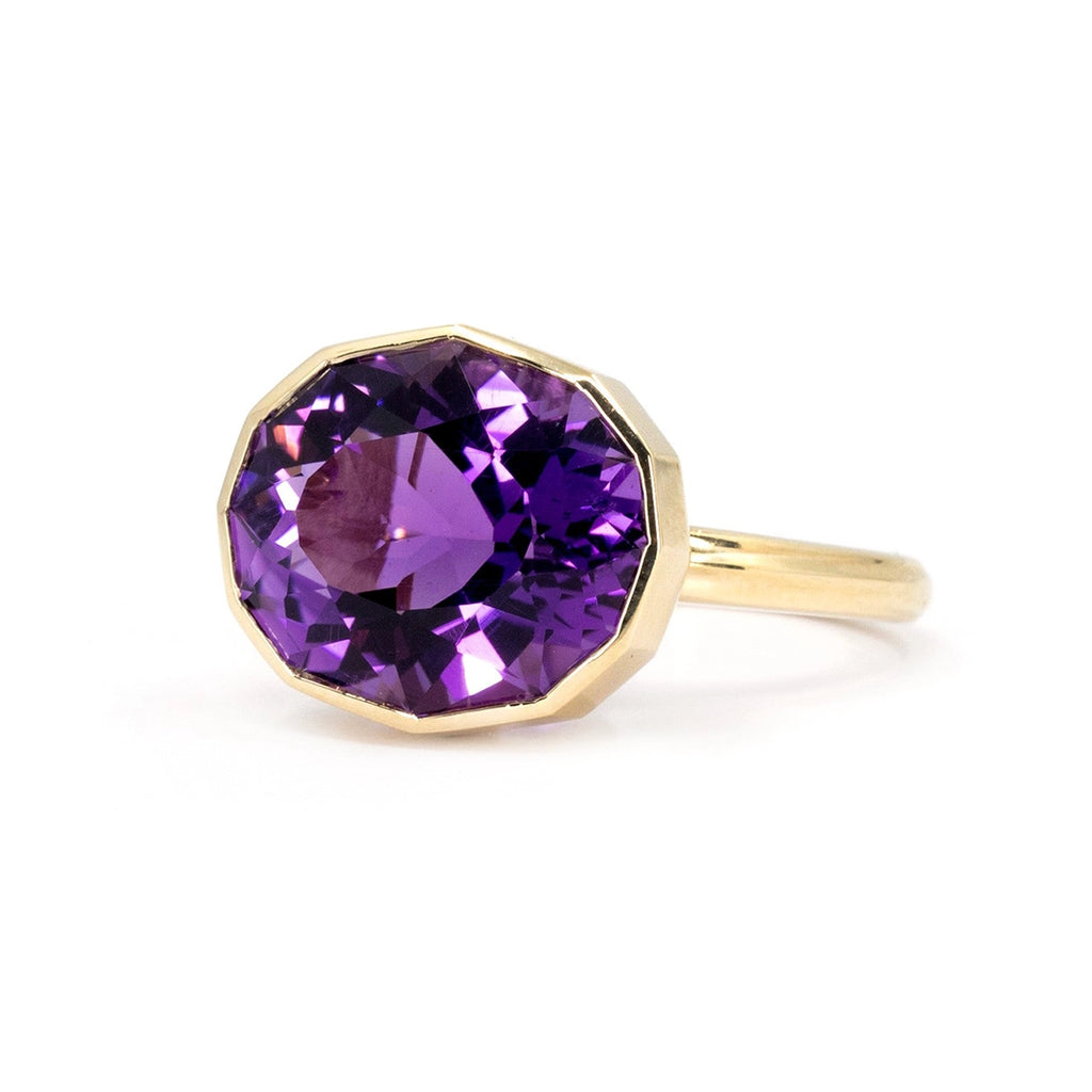 Side view of a big oval shape natural amethyst of profound purple color and pink hues bezel set in 14k yellow gold, seen photographed on a white background. This is a creation from Canadian jewelry designer Bena Jewelry. This unique piece of fine bold jewelry is available at Ruby Mardi.