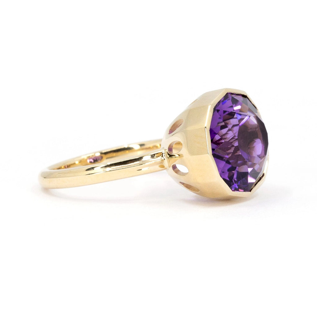 Side view of a big oval shape natural amethyst of profound purple color and pink hues bezel set in 14k yellow gold, seen photographed on a white background. We can see oval holes in the bold basket that let the light enter the gemstone. This is a creation from Montreal-based jewelry designer Bena Jewelry.