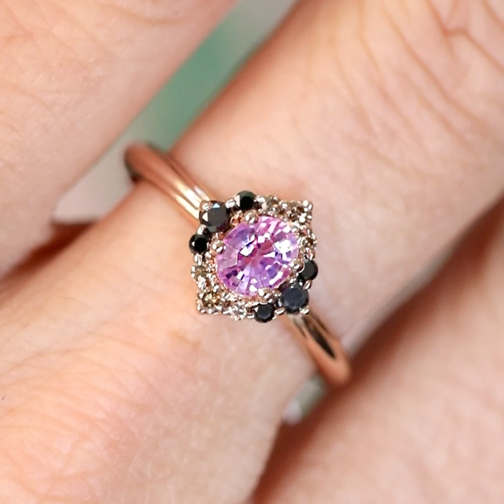 Velvet Aura ring worn on hand, featuring a vivid oval pink sapphire with black and champagne diamonds in a pointed rose gold halo. Ruby Mardi design.