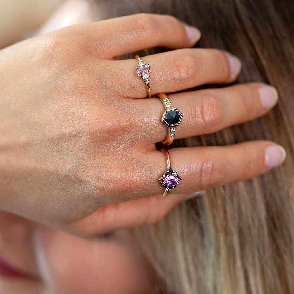 Close-up of a hand wearing three designer gemstone rings in rose gold: an oval pink sapphire engagement ring accented with diamonds, a bold hexagon black diamond ring with side stones, and a vintage-inspired pink sapphire ring framed by black diamonds. Elegant, colorful bridal and statement rings photographed worn for Ruby Mardi jewelry collection.