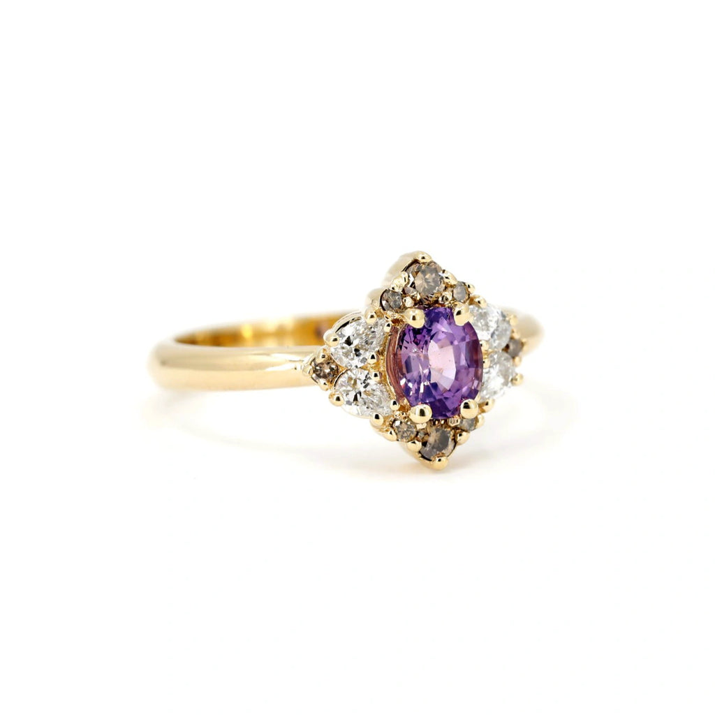 Angled view of a handcrafted yellow gold ring featuring an oval-cut purple sapphire at the center, surrounded by a halo of natural brown diamonds and brilliant white accent diamonds. The cluster design evokes a vintage yet modern feel, with asymmetrical sparkle and warm tones. Designed and made in Montreal by Ruby Mardi, this unique fine jewelry piece offers an artistic take on alternative engagement rings and bold gemstone settings.