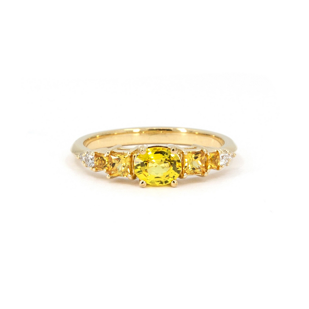 A product photography of an engagement ring in yellow gold with a central oval yellow sapphires and 4 side princess cut orange sapphires, as well as diamond accents. This fancy bridal ring was designed and handcrafted in Montreal, Canada, by Ruby Mardi.