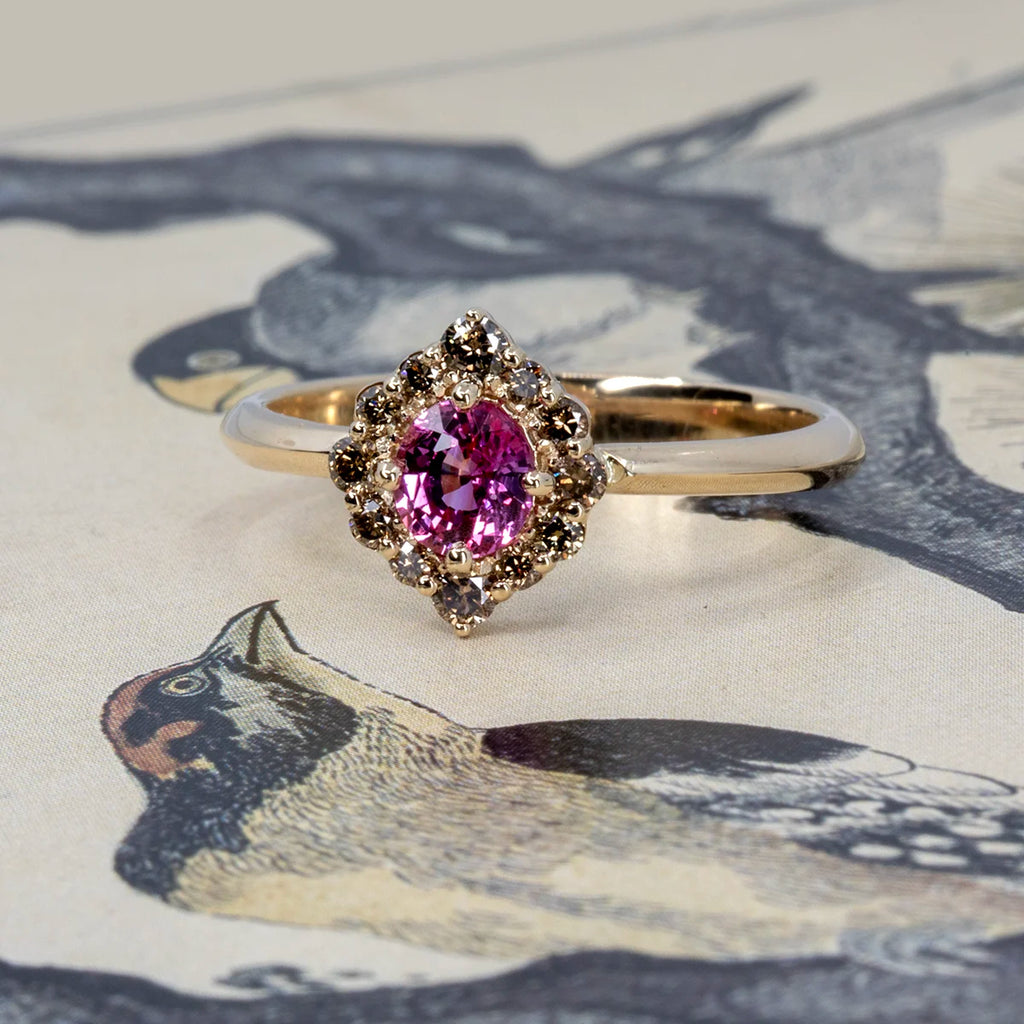 Yellow gold ring with a vivid pink sapphire surrounded by a halo of natural brown diamonds in a pointed floral setting. Photographed on an illustrated bird background. Handcrafted in Montreal by Ruby Mardi.