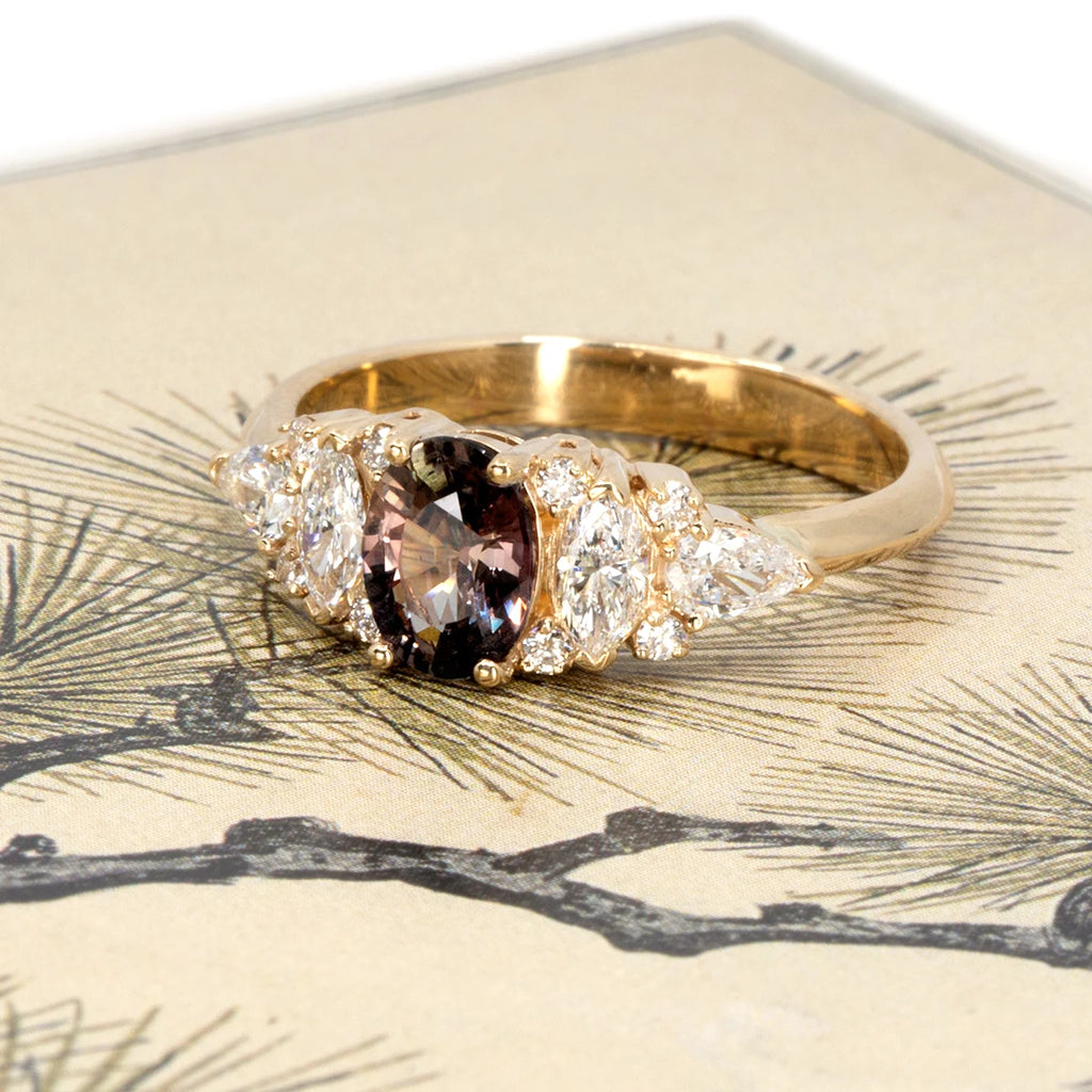 Yellow gold engagement ring with an oval plum sapphire at the center, flanked by marquise, pear, and round white diamonds. Photographed on a delicate botanical illustration background. Handcrafted by Ruby Mardi in Montreal.