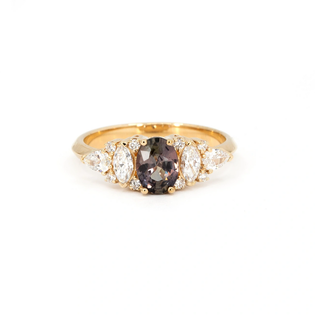 Plum sapphire engagement ring in yellow gold, featuring an oval-cut center stone with shifting hues of purple, bronze, and green, flanked by marquise, pear, and round white diamonds. Handcrafted in Montreal by Bena Jewelry, available at Ruby Mardi.