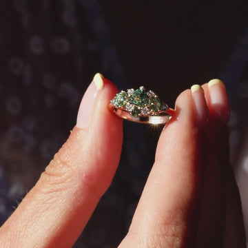 Close-up video of a rose gold cluster ring featuring green sapphires and white diamonds, held between two fingers and sparkling in natural sunlight.