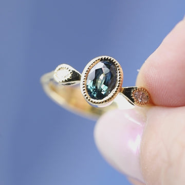 Short video showing an absolutely unique yellow gold ring filmed on a sky blue background. The band is formed by two superimposed snakes that join a central oval green sapphire on either side, set in a closed setting with miligrain detail. The gold snakes have white diamond eyes on the front and green diamond eyes on the sides! This exceptional engagement ring is available only at Canada's coolest jewelry store, Ruby Mardi. It was made by Emily Gill.