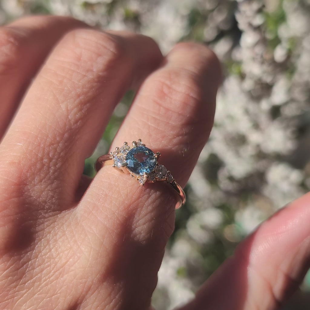 14 second video showing a one-of-a-kind engagement ring worn on an index. We can see the central navy blue sapphire and natural diamonds shine under the sun. The background of the video is out of focus, but we can guess it is white flowers. The bridal ring we see was handcrafted and designed by Justine Quintal, it is an asymmetrical ring with a round blue sapphire and cluster of white diamonds.
