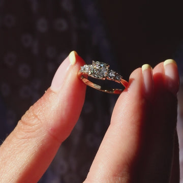 A hand wearing a 14k yellow gold engagement ring with an antique-cut cushion diamond. The diamond catches and reflects the sunlight as the hand gently moves, showcasing its sparkle and brilliance.