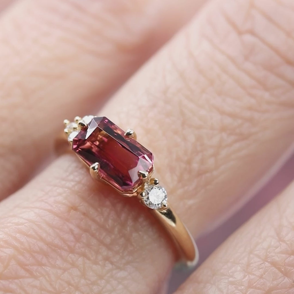 A video is showing a designer engagement ring in close up. The ring is made in yellow gold and features a big rectangular dark pink tourmaline as well as diamond accents. It's filmed on a blurry floral background. The creation is from indie designer Justine Quintal.