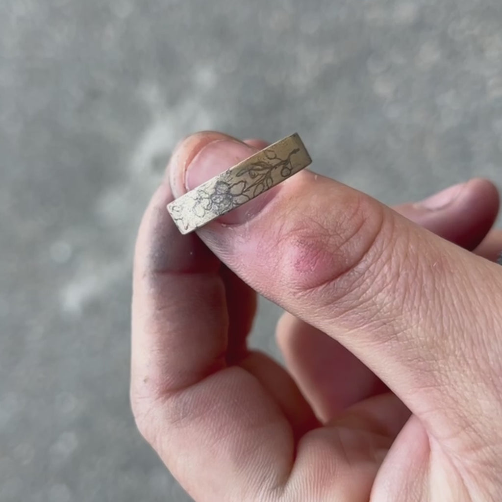 Video of a hand-engraved gold band held in the jeweller’s work-worn hands, showing the pencil-drawn floral design before engraving. The close-up captures the authentic, artisanal nature of the process, with visible bench dust and tool marks that highlight the craft behind Rebel & Rogue’s engraving work.