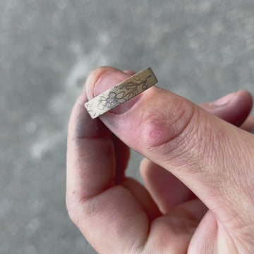 Video of a hand-engraved gold band held in the jeweller’s work-worn hands, showing the pencil-drawn floral design before engraving. The close-up captures the authentic, artisanal nature of the process, with visible bench dust and tool marks that highlight the craft behind Rebel & Rogue’s engraving work.