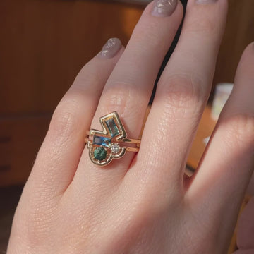 A video with an hawaïan music show a funky precious ring taking the sun light on a finger. The ring consists of a stunning geometric composition. A rounded yellow gold band encircles 4 gemstones: a white diamond, a round green sapphire, and two bicolored sapphire baguettes.