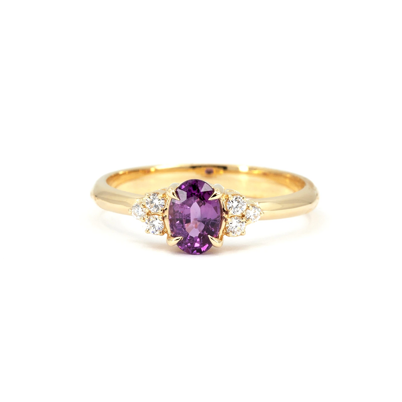  gold engagement ring featuring an oval-cut purple sapphire as the centerpiece, flanked by clusters of round brilliant-cut diamonds. The setting is crafted in yellow gold with delicate prongs holding the gemstones. The ring has a timeless and elegant design, blending vibrant color with classic sparkle. Photographed against a white background for a crisp and detailed view.