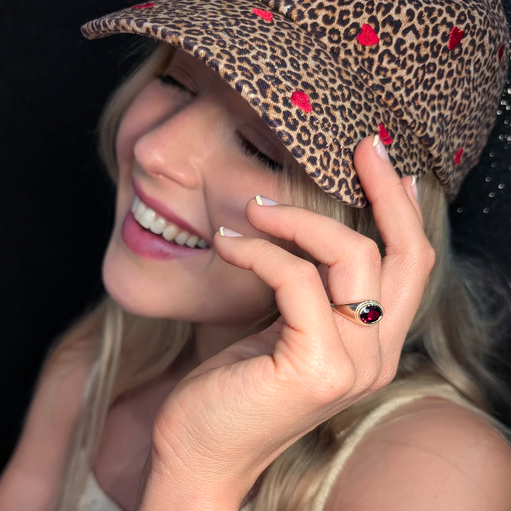 Smiling woman wearing a leopard-print cap with red hearts, showing a statement ring in 14k yellow gold set with a deep red pyrope garnet cabochon from Bena Jewelry Design, Montreal.