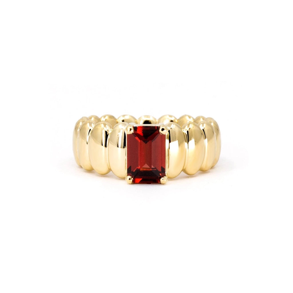 Bold yellow gold ring featuring a band made of elongated, rounded shapes, with a rectangular pyrope garnet in the center. This fashion ring with a dark red gemstone is made in Canada by the independent Bena Jewelry brand. This ring can be custom-ordered with your favorite stone. Shown here photographed against a white background.