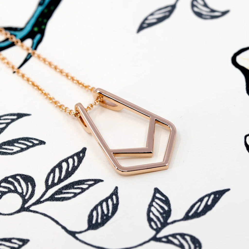 Chic Tie pendant by Bena Jewelry in rose gold, featuring a sleek geometric design. The pendant lies on an artistic black-and-white floral background, highlighting its polished finish. This modern statement piece doubles as a ring holder and is available at Ruby Mardi, a collective of independent designer jewelry in Canada.