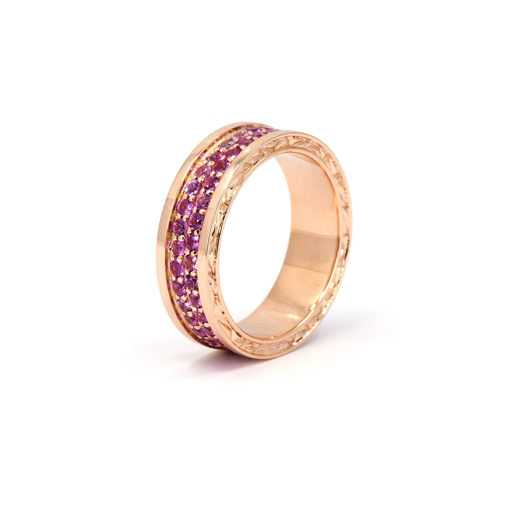 Angled view of a wide rose gold ring with two rows of vivid pink sapphires pavé-set in the center, framed by engraved sculptural edges. The intricate detailing on the sides adds depth and texture to the bold silhouette. Designed by Bena Jewelry and handcrafted in Montreal, this modern pink sapphire band is available exclusively at Ruby Mardi — a unique statement ring blending color, craftsmanship, and luxury.