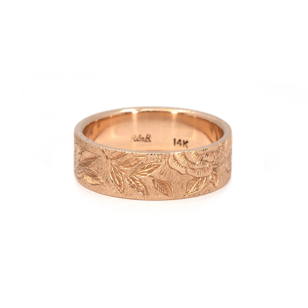 7 mm hand-engraved floral wedding band in 14k rose gold by Rebel & Rogue, featuring detailed peony and leaf engravings with a textured stippled background. Unique artisanal gold ring photographed for Ruby Mardi, Montreal jewelry store.