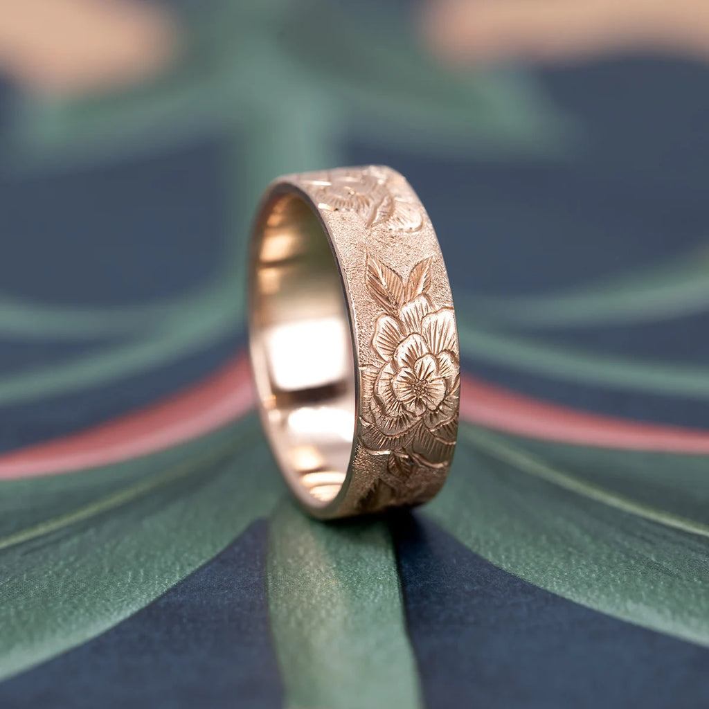 7 mm hand-engraved floral gold band by Rebel & Rogue, featuring detailed peony engravings with a stippled background. Solid yellow gold wedding ring photographed on a patterned surface at Ruby Mardi, Montreal jewelry store.