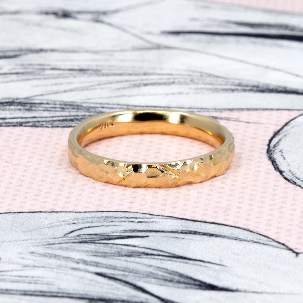 A hammered yellow gold wedding band with a rustic, textured finish, handcrafted by Sheena Purcell and displayed on a pink, sketch-like background—available at Ruby Mardi.