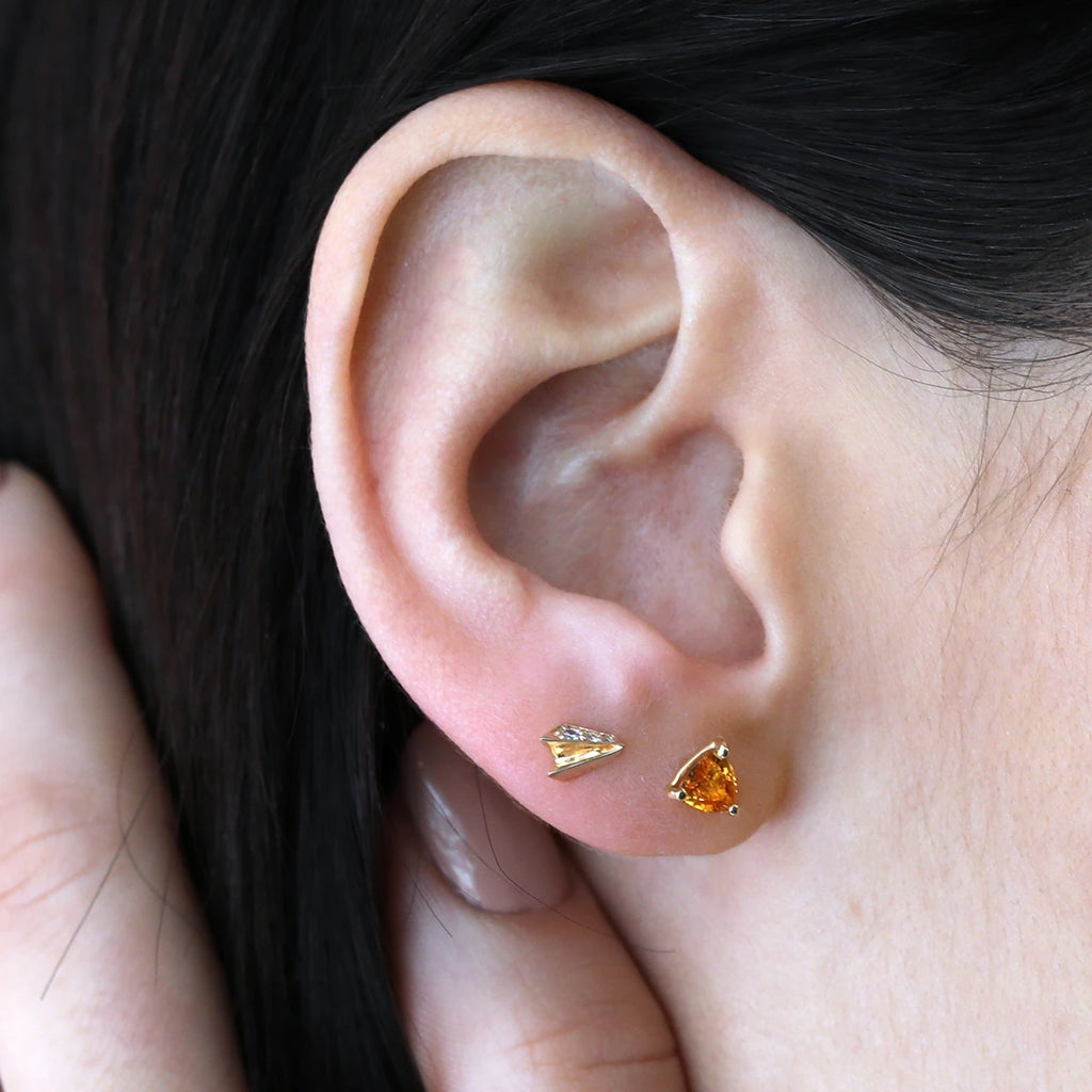 Close-up of a woman wearing two small, 18k yellow gold stud earrings: one with an orange sapphire and the other with diamonds, part of Bena Jewelry’s edgy collection. A striking yet subtle statement of modern elegance, photographed in natural light.