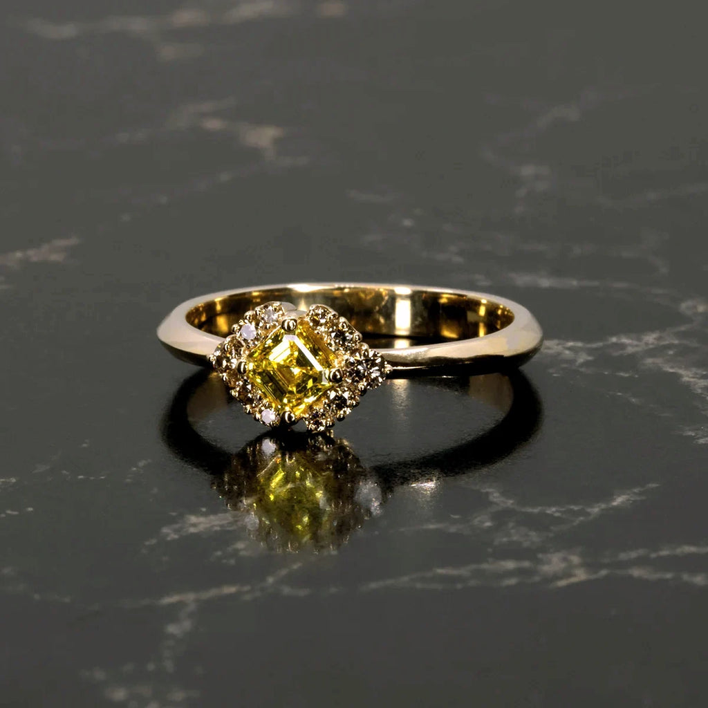 Yellow gold ring featuring an asscher-cut yellow diamond framed by champagne diamonds, displayed on a dark marble surface with a soft reflection.